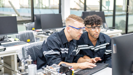 Two engineers sat at a computer in robot fabrication room quality checking electronic control board.