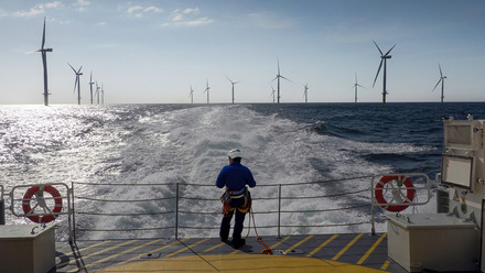 Rope access technician overlooks offshore wind farm and boat wake.